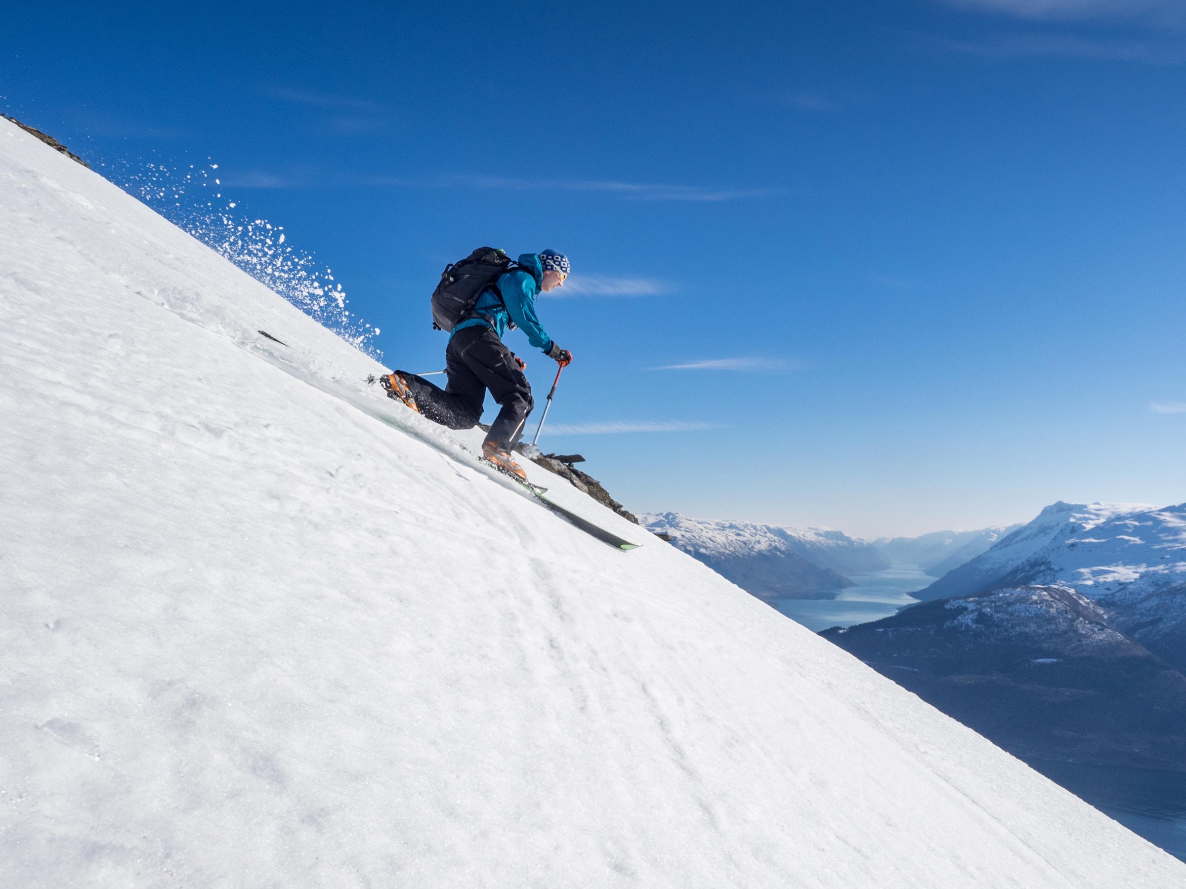 Jo Bjørnar på telemarkski i ein bratt snøbakke, med fjordar og fjell i bakgrunnen.
